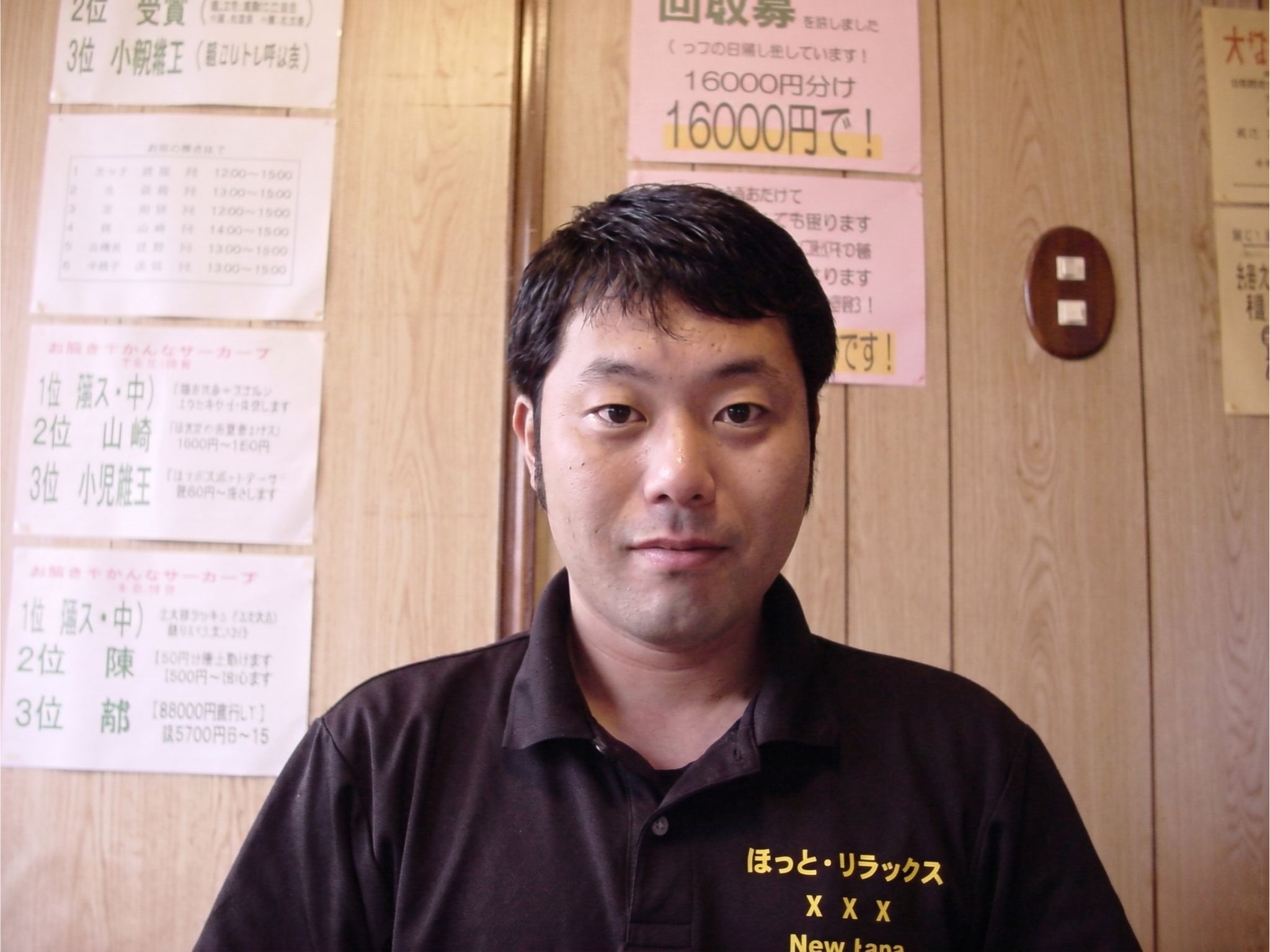沼津市の整体「ほっと、りらっくす整体」の代表、吉澤公亮のプロフィール写真です。黒いポロシャツのユニフォームを着た男性が、木目調の壁を背景に穏やかな表情で正面を向いており、当店をご利用になるお客様へ安心感をお伝えしています。